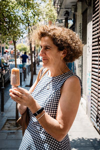 Mature woman with popsicle looking away while standing on sidewalk in city