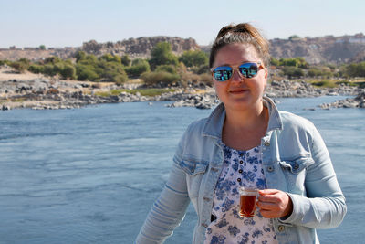 Portrait of woman in sunglasses having drink against lake