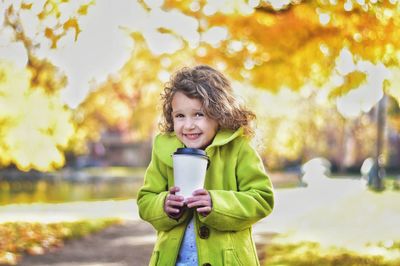 Portrait of cute smiling girl holding disposable cup while standing in park during autumn