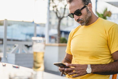 Mature man wearing sunglasses using mobile phone while sitting at sidewalk cafe