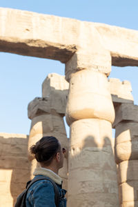 Vertical view of unrecognizable bun hair tourist young man looking up to the egyptian temple 