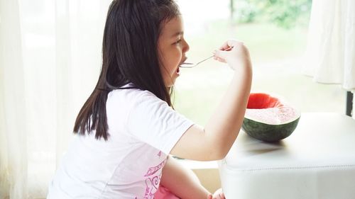 Side view of smiling girl having watermelon at home