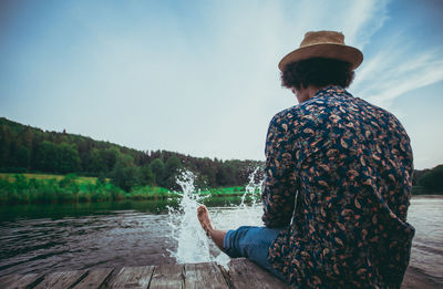 Rear view of man splashing water in lake while sitting on jetty against sky