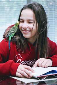 Teenage girl reading a book with green parakeet sitting on shoulder kissing