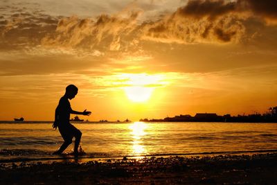 Silhouette boy standing at beach against sky during sunset