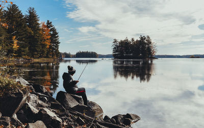 Full length of boy fishing while sitting on rock at lakeshore against cloudy sky