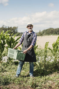 Portrait of smiling mature gardener holding crate and standing at farm