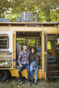 Portrait of smiling young multi-ethnic couple sitting at camper vehicle doorway during camping in forest