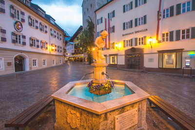 Illuminated fountain amidst buildings in city at dusk