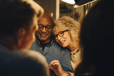 Smiling senior sharing smart phone with woman while sitting at illuminated dining table