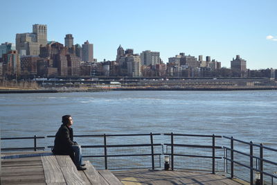 Woman sitting on railing by river against cityscape