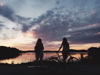 Silhouette female friends standing by lake against cloudy sky during sunset