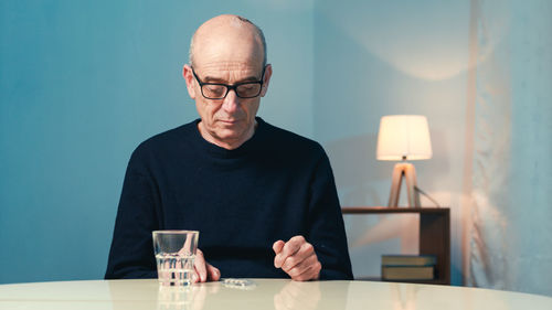 Elderly man with eyeglasses sitting at the table at home eats a heart pill