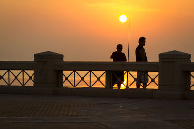 Silhouette of men standing by railing against sky during sunset