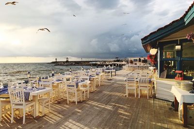 Empty chairs and tables at beach against sky