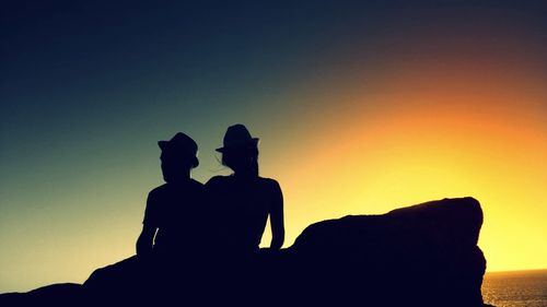 Silhouette couple sitting on rock by sea against sky during sunset
