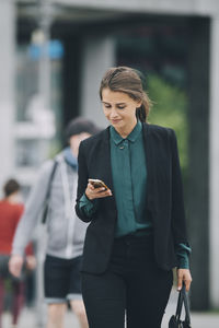 Confident businesswoman using smart phone while walking on street in city