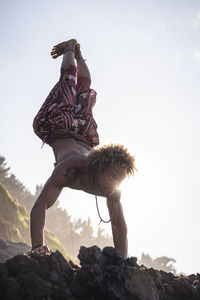Young man looking away while practicing handstand on rock at beach against sky