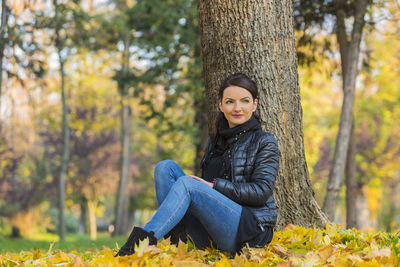 Thoughtful young woman sitting against tree trunk at park during autumn