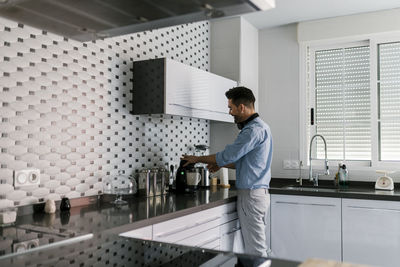 Man with headphones using coffee maker while standing in kitchen at home