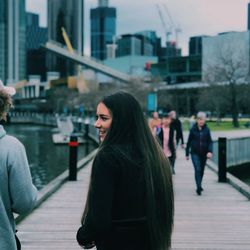 Rear view of smiling young woman standing on boardwalk in city