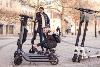 Teenager unlocking electric push scooter with smart phone while crouching by friend on street in city