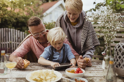 Younger brother using mobile phone sitting by father and sibling at table in dinner party