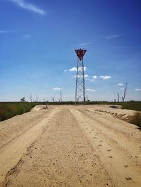 Dirt road with tire tracks leading towards metal tower against sky