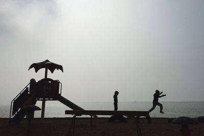 Silhouette boys playing at playground by sea against sky