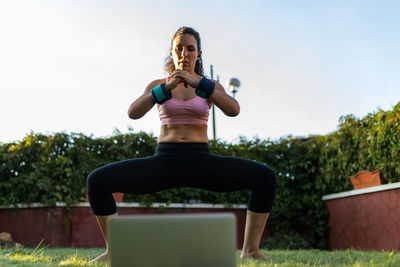 From below fit female athlete in sportswear using laptop and doing squats while following online tutorial during training in backyard at sunset