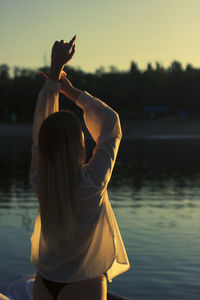 A young woman stands with her back to the camera near water at sunset with her hands up