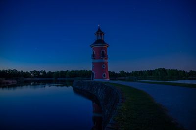 Lighthouse by lake and buildings against sky at dusk