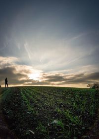 Distant view of silhouette man standing on grassy field against sky during sunset