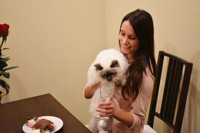 Happy woman with cat holding glass while sitting on chair at home