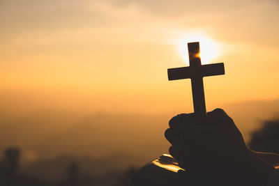 Silhouette person holding cross against sky during sunset