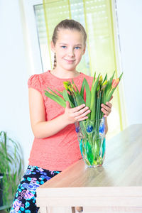 Portrait of teenage girl holding flowers in vase on wooden table at home