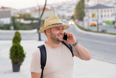 Handsome bearded smiling man,tourist, wearing sunhat, backpack, having call using smartphone