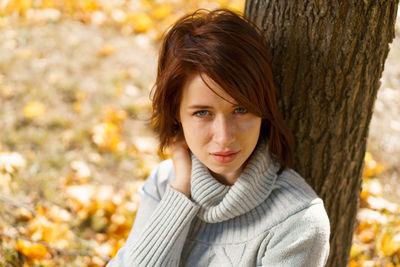 A young woman in a gray sweater sits alone, leaning against a tree