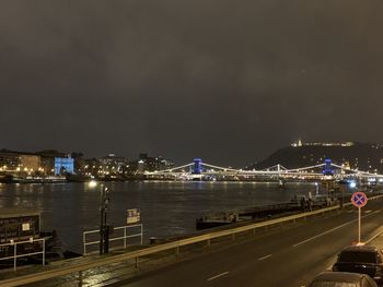 Illuminated bridge over river by buildings against sky at night