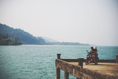People sitting on pier looking at lake against sky