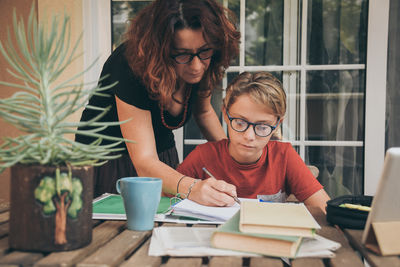 Young student doing homework at home with school books, and digital pad helped by his mother