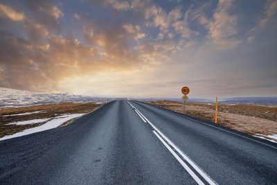 Diminishing road amidst snow covered landscape against cloudy sky during sunset