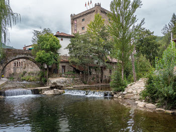 Bridge over river amidst trees and buildings against sky