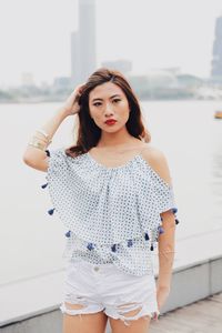 Portrait of young woman standing on footpath against river in city