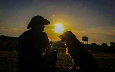 Rear view of man sitting with dog on field against sky during sunset