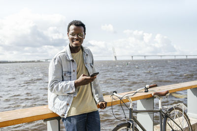 Younh´g man leaning on railing by the sea, using smartphone