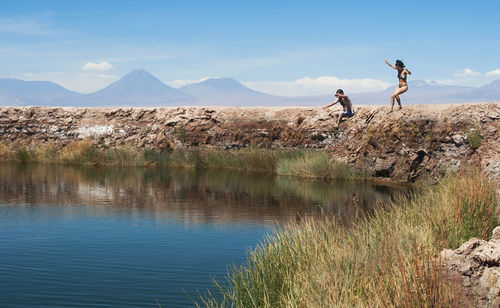 Scenic view of mountains against sky and a young man and woman jumping to a lake 