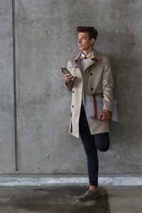 Young model holding mobile phone and bag while leaning on wall