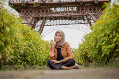 Beautiful asian woman in hijab sitting with hand on chin in flower garden path