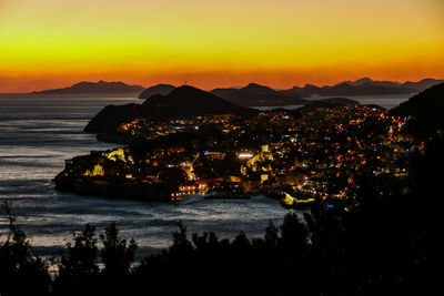 Illuminated city by sea against sky during sunset